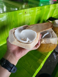 A freshly halved young coconut held in a person's hand, revealing the soft, white flesh and juice inside. A green, textured table surface provides a vibrant background, with another coconut and straw partially visible on the side.