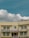 A two-story beige building with multiple windows and a prominent sign in red text in the middle, flanked by some smaller signs and flags. A satellite dish is mounted on the roof, and electrical cables run across the front of the building. The sky is clear with some fluffy white clouds.