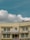 A two-story beige building with multiple windows and a prominent sign in red text in the middle, flanked by some smaller signs and flags. A satellite dish is mounted on the roof, and electrical cables run across the front of the building. The sky is clear with some fluffy white clouds.