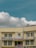 A two-story beige building with multiple windows and a prominent sign in red text in the middle, flanked by some smaller signs and flags. A satellite dish is mounted on the roof, and electrical cables run across the front of the building. The sky is clear with some fluffy white clouds.