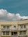 A two-story beige building with multiple windows and a prominent sign in red text in the middle, flanked by some smaller signs and flags. A satellite dish is mounted on the roof, and electrical cables run across the front of the building. The sky is clear with some fluffy white clouds.