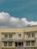 A two-story beige building with multiple windows and a prominent sign in red text in the middle, flanked by some smaller signs and flags. A satellite dish is mounted on the roof, and electrical cables run across the front of the building. The sky is clear with some fluffy white clouds.