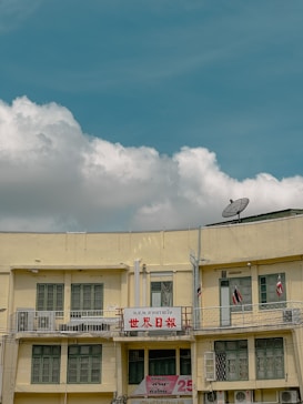 A two-story beige building with multiple windows and a prominent sign in red text in the middle, flanked by some smaller signs and flags. A satellite dish is mounted on the roof, and electrical cables run across the front of the building. The sky is clear with some fluffy white clouds.