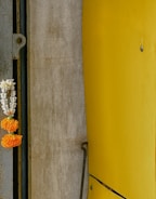 A vertical composition featuring a textured gray concrete wall on the left, adorned with a hanging floral garland made of orange marigolds and white flowers. Adjacent to it, on the right, a smooth, bright yellow curved metallic surface with visible fasteners.