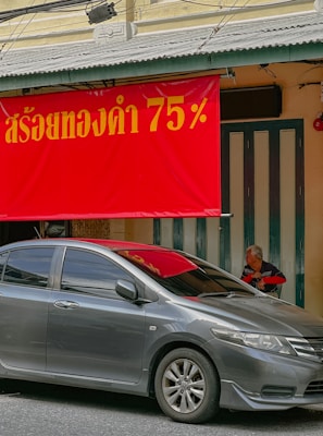 A gray car is parked on the street in front of a building. Above the car is a large red sign with yellow text. An elderly person is sitting in the doorway behind the car.