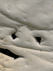 A close-up view of a white fabric with several irregularly shaped holes, frayed edges, and a slightly wrinkled texture. The fabric appears to be old or worn out, with visible discoloration and wear.