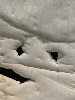 A close-up view of a white fabric with several irregularly shaped holes, frayed edges, and a slightly wrinkled texture. The fabric appears to be old or worn out, with visible discoloration and wear.