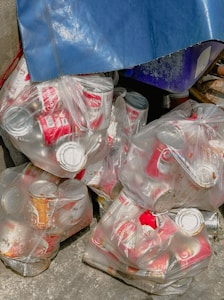 Plastic bags filled with used red and white aluminum cans are piled together. There is a blue tarp covering part of the pile and the ground is made of concrete.