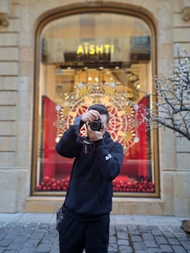 A person stands on a cobblestone street holding a camera, taking a photo. They are wearing a black hoodie and are positioned in front of a store window with the word 'AÏSHTI' visible. The window display features a decorative circular pattern and red accents, possibly holiday themed. A tree with bare branches is partially visible on the right.