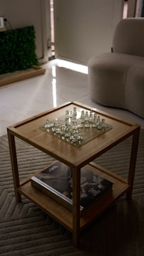 A wooden table with a glass chess set on top and a large book on the lower shelf. The room includes a textured gray carpet, a light-colored armchair, and a partial view of greenery by a wall.