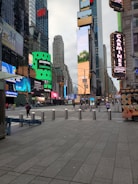 A bustling urban scene featuring tall skyscrapers adorned with numerous bright and colorful digital billboards. The area appears to be a well-known city square with commercial spaces and advertisements. People are walking on the wide pedestrian paths, and there are bollards lining the street for safety. To the right, there's a street vendor cart with images of food.
