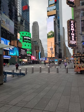 A bustling urban scene featuring tall skyscrapers adorned with numerous bright and colorful digital billboards. The area appears to be a well-known city square with commercial spaces and advertisements. People are walking on the wide pedestrian paths, and there are bollards lining the street for safety. To the right, there's a street vendor cart with images of food.