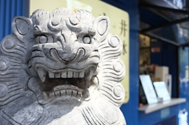 A stone lion sculpture with intricate carvings stands prominently in a detailed close-up, showing its fierce and majestic facial features. The background includes a partially blurred storefront with signage and modern elements.
