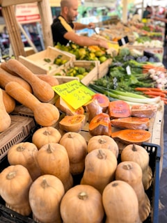 A bustling market stall displays a variety of fresh vegetables, prominently featuring squash and pumpkins. Other produce includes carrots, leafy greens, and zucchini. A vendor stands behind the counter, assisting customers.