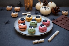 A plate of colorful macarons with playful monster faces complete with sugar eyes and decorations resembling black flowers. Surrounding the plate are decorative items, including white pumpkins, pine cones, small black spiders, and a brown silicone mat with spider designs. Near the plate, lettered cubes spell out 'HAPPY HALLOWEEN'. The scene is set for a Halloween-themed arrangement with a cozy, whimsical feel.