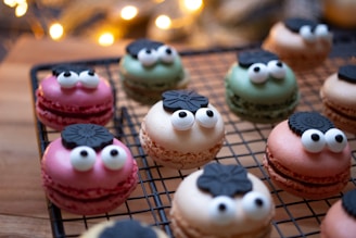 A display of colorful macarons designed with expressive eyes and decorative chocolate tops, resting on a cooling rack. The macarons are in shades of pink, green, and beige, creating a playful and festive atmosphere with blurred warm lights in the background.