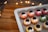 A tray filled with colorful macarons decorated with googly eyes and dark toppings resembling flowers. The macarons are arranged in rows, showcasing a variety of colors including pink, orange, and green. In the background, there's a warm ambiance created by softly glowing string lights.