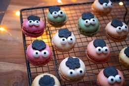 A collection of colorful macarons with playful googly eyes and black flower-shaped decorations, arranged on a cooling rack. The background features warm string lights and a wooden surface, creating a cozy atmosphere.