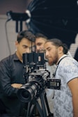Three people are gathered around a professional video camera setup, appearing focused and engaged. The camera is large and mounted on a tripod, with various controls and attachments visible. The background has a soft focus, indicating a studio setting possibly used for film or photography.