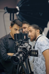 Three people are gathered around a professional video camera setup, appearing focused and engaged. The camera is large and mounted on a tripod, with various controls and attachments visible. The background has a soft focus, indicating a studio setting possibly used for film or photography.