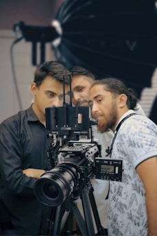 Three people are gathered around a professional video camera setup, appearing focused and engaged. The camera is large and mounted on a tripod, with various controls and attachments visible. The background has a soft focus, indicating a studio setting possibly used for film or photography.