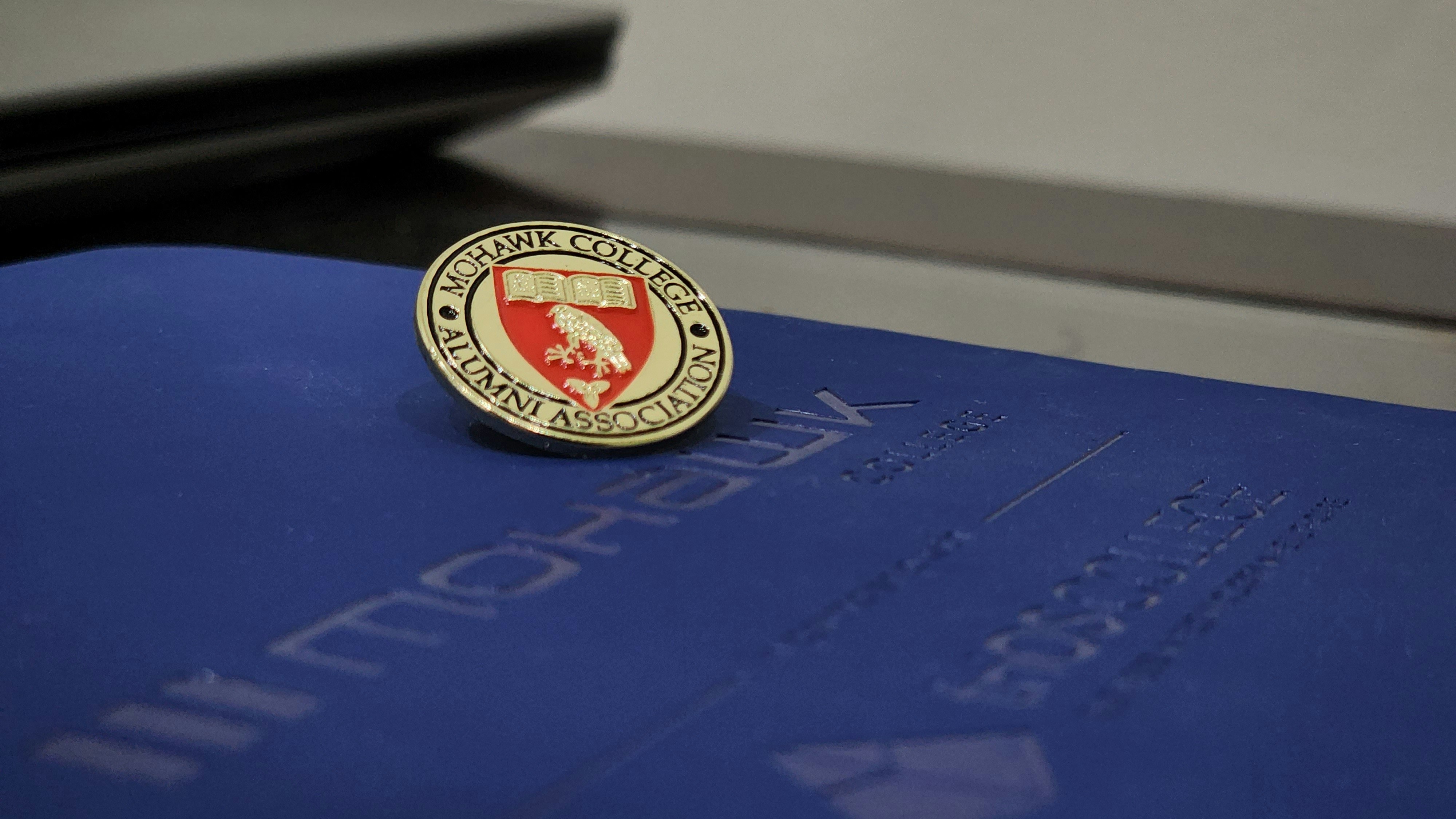 A Mohawk College Alumni Association pin is placed on top of a dark blue folder or booklet. The pin features a red background with an engraved book and branch symbol, surrounded by the words 'Mohawk College Alumni Association' in gold lettering.
