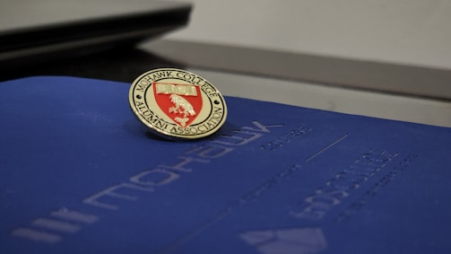 A Mohawk College Alumni Association pin is placed on top of a dark blue folder or booklet. The pin features a red background with an engraved book and branch symbol, surrounded by the words 'Mohawk College Alumni Association' in gold lettering.
