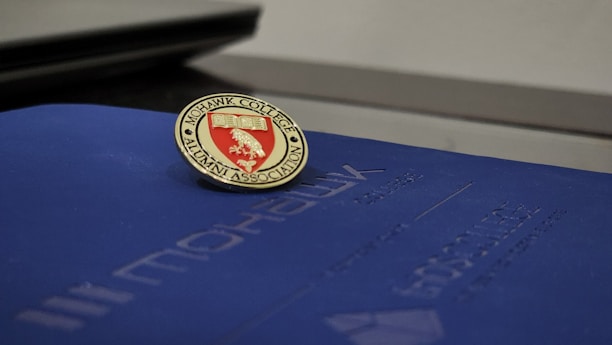 A Mohawk College Alumni Association pin is placed on top of a dark blue folder or booklet. The pin features a red background with an engraved book and branch symbol, surrounded by the words 'Mohawk College Alumni Association' in gold lettering.