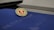 A Mohawk College Alumni Association pin is placed on top of a dark blue folder or booklet. The pin features a red background with an engraved book and branch symbol, surrounded by the words 'Mohawk College Alumni Association' in gold lettering.
