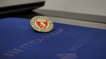 A Mohawk College Alumni Association pin is placed on top of a dark blue folder or booklet. The pin features a red background with an engraved book and branch symbol, surrounded by the words 'Mohawk College Alumni Association' in gold lettering.