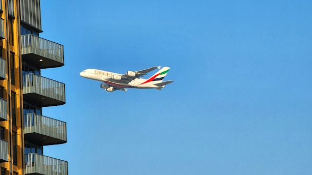 A large commercial airplane with the Emirates logo is flying against a clear blue sky near a modern residential building. The building has several balconies with yellow external walls and gray railings.
