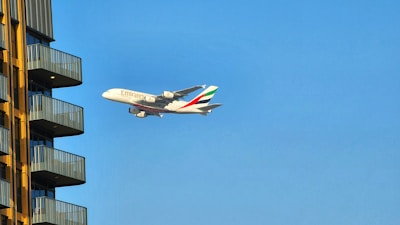 A large commercial airplane with the Emirates logo is flying against a clear blue sky near a modern residential building. The building has several balconies with yellow external walls and gray railings.