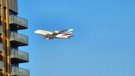 A large commercial airplane with the Emirates logo is flying against a clear blue sky near a modern residential building. The building has several balconies with yellow external walls and gray railings.