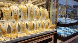 A showcase displaying a collection of intricate gold jewelry, including necklaces, earrings, bracelets, and rings, arranged on white busts and stands. The gold jewelry is highly ornate and detailed, with different designs and patterns. In the background, additional jewelry pieces are visible in a separate section behind glass.