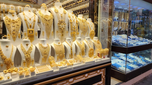 A showcase displaying a collection of intricate gold jewelry, including necklaces, earrings, bracelets, and rings, arranged on white busts and stands. The gold jewelry is highly ornate and detailed, with different designs and patterns. In the background, additional jewelry pieces are visible in a separate section behind glass.