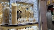 A jewelry store window display filled with mannequins adorned in elaborate, gold jewelry sets. The items include necklaces, bracelets, and full body chains, all intricately designed. The backdrop features a tufted, possibly velvet texture, adding to the luxurious atmosphere.