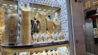 A jewelry store window display filled with mannequins adorned in elaborate, gold jewelry sets. The items include necklaces, bracelets, and full body chains, all intricately designed. The backdrop features a tufted, possibly velvet texture, adding to the luxurious atmosphere.