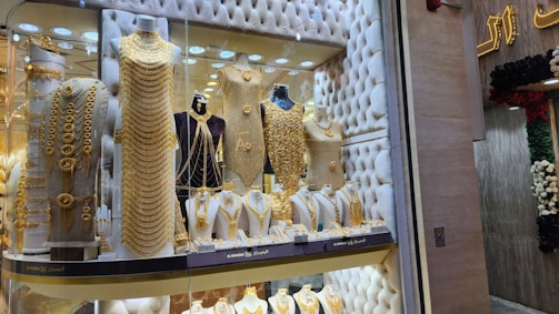 A jewelry store window display filled with mannequins adorned in elaborate, gold jewelry sets. The items include necklaces, bracelets, and full body chains, all intricately designed. The backdrop features a tufted, possibly velvet texture, adding to the luxurious atmosphere.