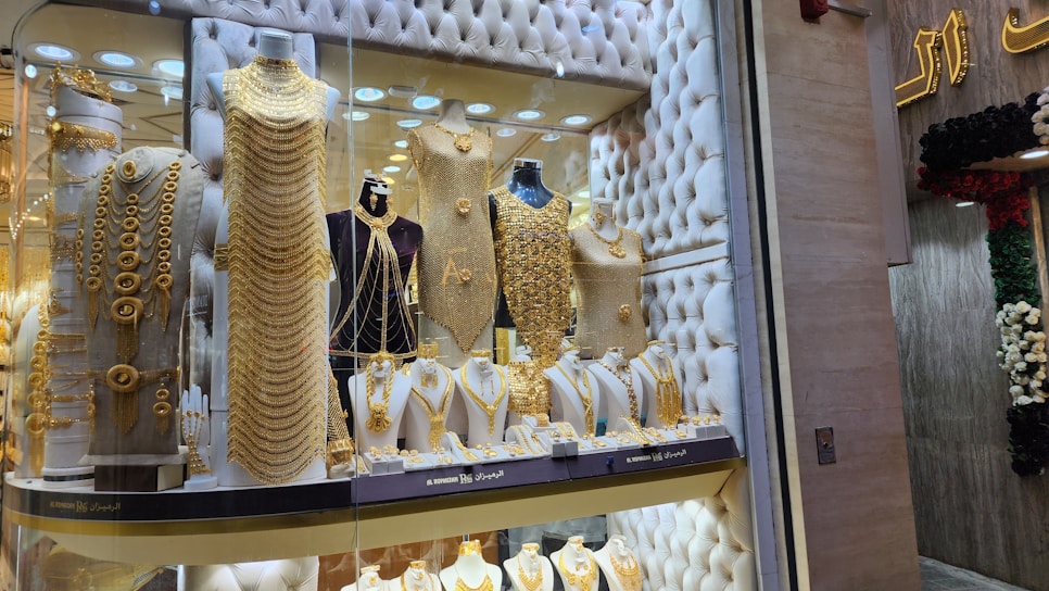A jewelry store window display filled with mannequins adorned in elaborate, gold jewelry sets. The items include necklaces, bracelets, and full body chains, all intricately designed. The backdrop features a tufted, possibly velvet texture, adding to the luxurious atmosphere.