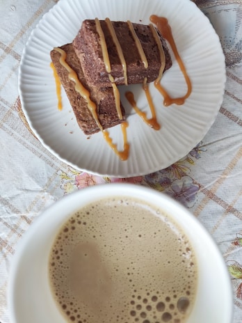 Two caramel-drizzled brownies are placed on a white plate with a textured rim, set on a table with a floral and plaid-patterned tablecloth. Below the plate is a cup filled with frothy coffee, visible at the bottom of the image.