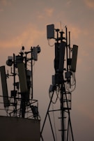 Several communication towers with antennas and dishes are silhouetted against a dusky sky. The metal structures are adorned with various electronic components and cables. The soft light from the setting sun creates a warm ambiance in the background.