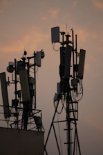 Several communication towers with antennas and dishes are silhouetted against a dusky sky. The metal structures are adorned with various electronic components and cables. The soft light from the setting sun creates a warm ambiance in the background.