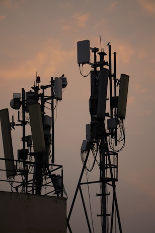 Several communication towers with antennas and dishes are silhouetted against a dusky sky. The metal structures are adorned with various electronic components and cables. The soft light from the setting sun creates a warm ambiance in the background.