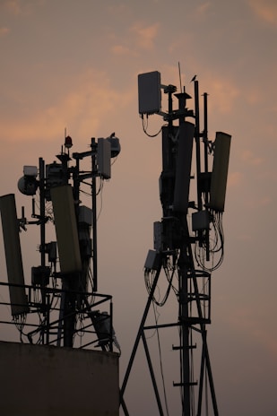 Several communication towers with antennas and dishes are silhouetted against a dusky sky. The metal structures are adorned with various electronic components and cables. The soft light from the setting sun creates a warm ambiance in the background.