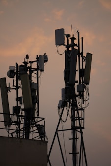 Several communication towers with antennas and dishes are silhouetted against a dusky sky. The metal structures are adorned with various electronic components and cables. The soft light from the setting sun creates a warm ambiance in the background.