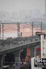 An elevated metro railway track runs across the frame with metal support structures and wires. Below, a busy street scene includes vehicles and billboards in an urban environment. In the background, numerous high-rise buildings are visible through a layer of haze, suggesting a metropolitan area.