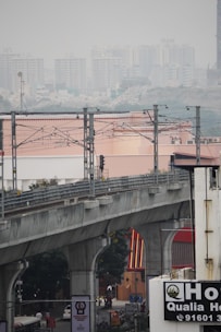 An elevated metro railway track runs across the frame with metal support structures and wires. Below, a busy street scene includes vehicles and billboards in an urban environment. In the background, numerous high-rise buildings are visible through a layer of haze, suggesting a metropolitan area.