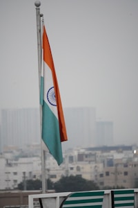 An Indian national flag is flying on a pole against a backdrop of an overcast sky and a blurred cityscape in the background. The flag has three horizontal stripes of orange, white, and green, with a blue Ashoka Chakra in the center.