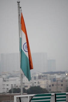 An Indian national flag is flying on a pole against a backdrop of an overcast sky and a blurred cityscape in the background. The flag has three horizontal stripes of orange, white, and green, with a blue Ashoka Chakra in the center.