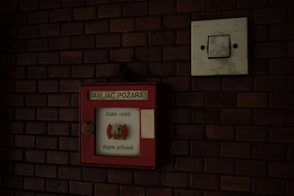 A red fire alarm box is mounted on a dark brick wall. The box has a label with text written in a foreign language, possibly indicating instructions or information for emergency situations. Next to the box is a square, white electrical switch panel.